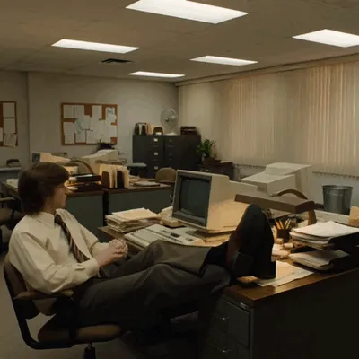 With his feet on the desk, a man turns a wastebasket into a basketball hoop.