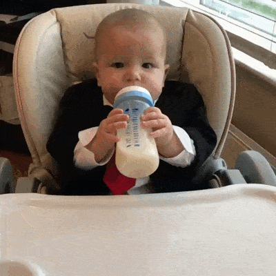Un bébé en costume et cravate rouge boit son biberon dans une chaise haute.