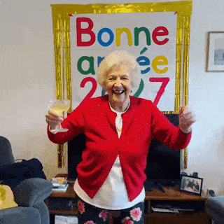 Une mamie en veste rouge danse et lève son verre devant une affiche Bonne année 2027.