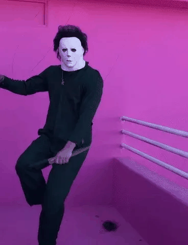 Um homem com roupa preta e máscara branca dança em um quarto rosa.