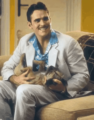 Matt Dillon usa um sorriso forçado com um cachorro pequeno no colo.