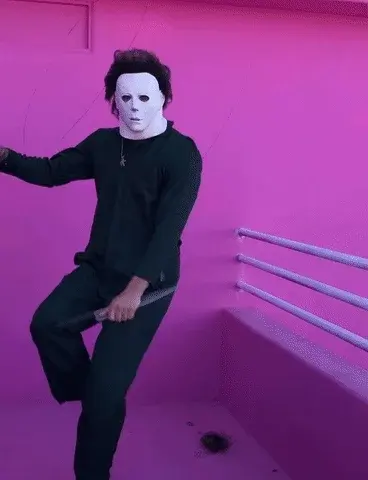 Un homme habillé en noir, portant un masque blanc, danse dans une pièce rose.