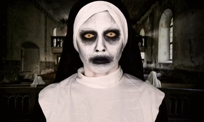 A pale-faced nun with yellow eyes and an unsettling stare stands in a dark church.