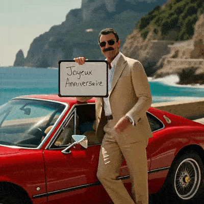 Un homme tient une ardoise Joyeux Anniversaire près d’une voiture rouge et de la mer.