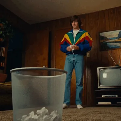 In a retro living room, a man in a colorful jacket aims a paper ball at a wastebasket.
