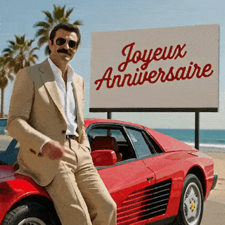 Magnum pointe vers la caméra près d’une voiture rouge et d’un panneau blanc Joyeux Anniversaire.