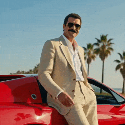 Magnum pose près d’une voiture rouge et d’un panneau bleu Joyeux Anniversaire.