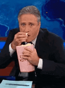 Un homme en costume mange du popcorn dans une boîte rayée, assis à un bureau devant un décor bleu.