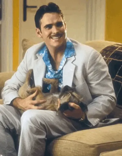 Matt Dillon wears a strained smile with a small dog on his lap.