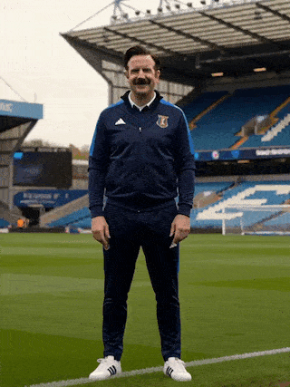 Ted Lasso pointe vers la caméra en souriant sur un terrain de football.